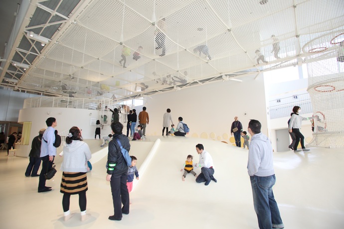 白を基調とした広々とした屋内遊戯施設で、天井に張り巡らされた巨大なネット遊具の上や、その下の白いドーム状の滑り台でたくさんの子どもたちが遊び、その様子を保護者たちが見守っている写真