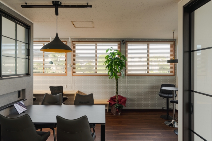 ブラインドの付いた窓から光が差し込む落ち着いたオフィス空間に、黒いデスクやチェア、観葉植物が配置され、手前には黄色い裏地のペンダントライトが吊るされたモダンな部屋の写真