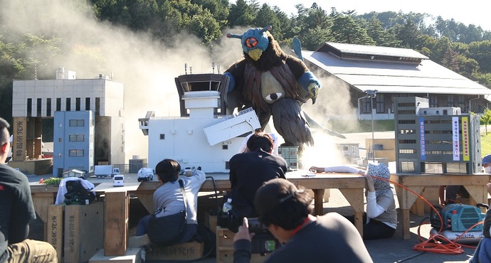 ミニチュアの町のセットの中で、鳥のくちばしのようなものが付いた怪獣が歩いている様子を数名の男性が撮影している、特撮塾の様子の写真
