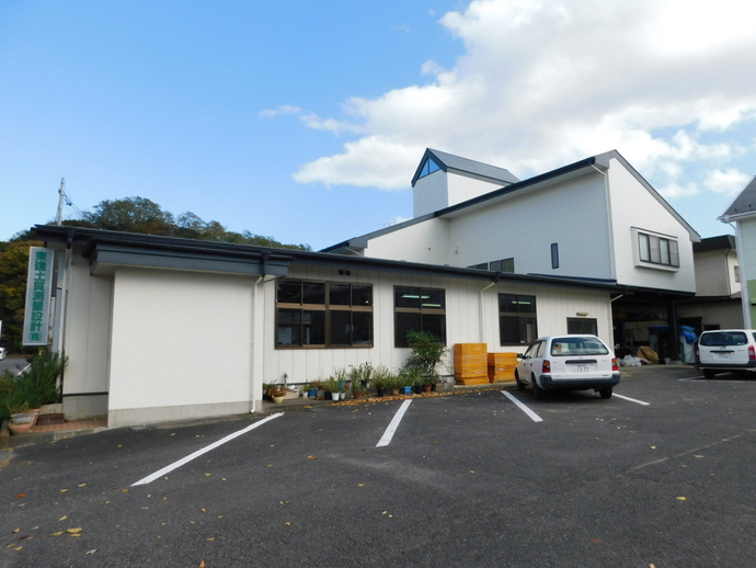 青空の下、白い外壁が特徴的な平屋建ての事務所ビルの前に、数台の乗用車が停車している広々としたアスファルトの駐車場がある東建土質測量設計株式会社の外観写真