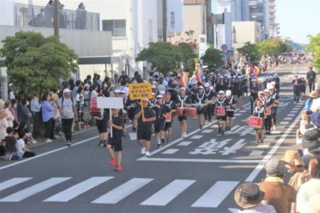 市街地の道路で行われているパレードの様子で、プラカートを持った先頭の2人に続き、黒のユニフォームを着た鼓笛隊が並んで歩き、沿道では大勢の人々が見物をしている様子の写真