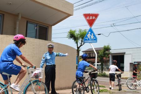 建物の角にある交差点で、青い制服を着た男性が腕を伸ばして指示を出し、ヘルメットを着用した子どもたちが、「止まれ」の標識の前で自転車を降り、自転車を押して歩いている様子の写真