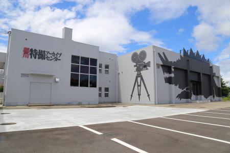 灰色の外壁の建物で、左側には「須賀川特撮アーカイブセンター」と赤と黒の文字、右側の壁には大型の映画撮影用カメラと怪獣のシルエットが黒で描かれており、前面には広い舗装駐車場がある施設の外観写真