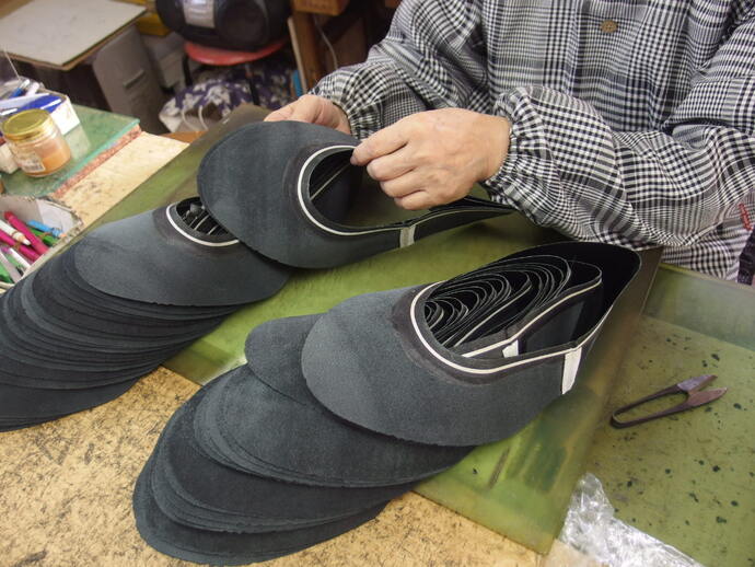 チェック柄の上衣を着た人物が、革靴またはスリッパの甲の部分に使うと思われる黒いフェルトのような生地の部品を、台の上で重ねながら加工している作業風景の写真