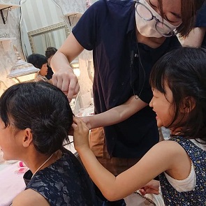 室内で椅子に座る人物の髪を整えている様子が写され、別の人物が横から髪を持って手伝っている場面で、背景には鏡やライトが並ぶヘアメイクアップアーティスト体験風景の写真