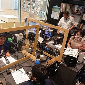 アクリルパネルが設置されたラジオブース内で、マイクに向かって原稿を読む数名の男女と、それを見守るように背後に立つ男性が見えるラジオパーソナリティ体験の風景写真