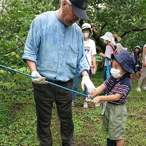 果樹園のような緑の木々に囲まれた屋外で、長袖シャツと帽子を着用した人物が手袋をして収穫用の長い棒を持ち、隣で子どもがその棒を握って作業を手伝っている農業体験の様子が写された写真