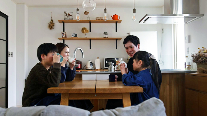木目調の明るいキッチンの前に置かれた木製のダイニングテーブルを囲んで、家族4人が飲み物を手に、楽し気に話をしている様子の写真