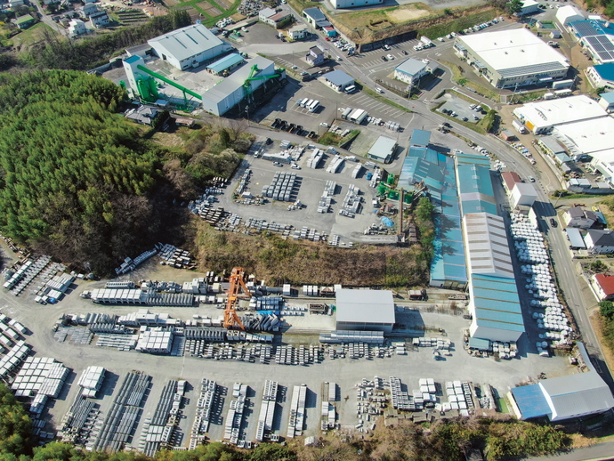 広大な敷地に、水色やグレーの屋根の複数の大きな建物が建ち、コンクリート製の資材が整然と積まれている様子の空撮写真