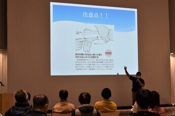 前方の大きなスクリーンに「注意点！！」と題された資料が映し出され、その前に立った講師の男性が資料を指して説明をしている様子の写真