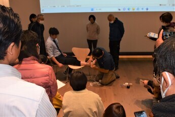 大きなスクリーンの前に置かれた椅子に座った男性が、もう一つの椅子に片足を伸ばして乗せ、講師の男性が足首にテーピングを行っている様子を、周りに集まった参加者たちが熱心に見学している様子の写真