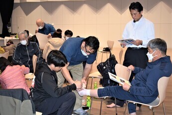 二人一組でテーピングの実技演習を行っている参加者が講師の男性からの指導を受けており、後方で資料を持った男性が見守っている様子の写真