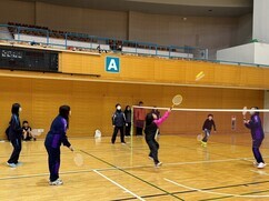 体育館にて、ラケットを高く掲げた選手がネット越しに鋭いショットを放ち、それに応じる対戦相手や周囲で見守る参加者たちがシャトルの行方を真剣に追いかけている、活気あるバドミントン競技の試合風景写真