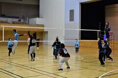 体育館にて、ネットを挟んで水色のシャツを着たチームと黒いユニフォームのチームが向かい合い、アタッカーが跳躍してスパイクを放つ瞬間を他の選手たちが身構えて見守っている、ラバーバレーボール試合の白熱した風景写真