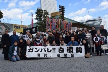 青空の下で「ガンバレ!! 白熊関須賀川後援会」と書かれた大きな横断幕を広げ、力士の四股名が入った旗やうちわを手にした大勢の支援者たちが、両国国技館を背景に笑顔で並んで応援の意気込みを示す集合写真