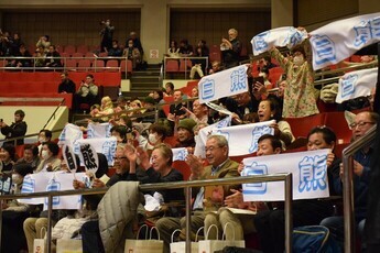 大相撲の観客席にて、大勢の支援者たちが「白熊」と青い文字で書かれた応援タオルを広げたり、うちわを振ったりしながら、笑顔や拍手で熱心に声援を送っている、会場が一体となった活気あふれる応援風景の写真