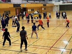 体育館にてオレンジ色のシャツを着た三人の選手がバドミントンの試合を行っており、手前のコートで待機する三人の選手や審判、周囲で見守る多くの観客が木目調のフロアに集まっているバトミントン大会の写真