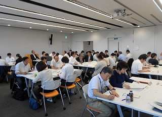 広い会議室に複数の四角いテーブルが並び、大勢の参加者がテーブルを囲んで座り、資料を広げて協議を行っている様子の写真