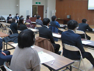 会議室で、たくさんの参加者が机に並んで座り、前方のスクリーンに注目している様子を後方から写した写真