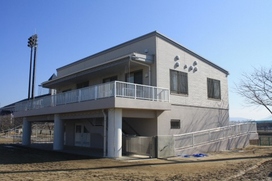 青空の下、グラウンドの脇に建てられた2階建てのスポーツ施設で、1階部分が吹き抜けのピロティ構造、2階には白い手すりのある広いベランダが備わっている建物を斜め前から捉えた写真