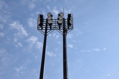 抜けるような青空に向かって、2本の高い支柱の先端に合計12個の円形ライトが格子状に整然と並び、スポーツ施設やグラウンドを照らすための巨大な照明塔を見上げるように捉えた写真