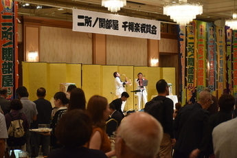 「二所ノ関部屋千秋楽祝賀会」と書かれた横断幕が掲げられた華やかな会場で、金屏風を背にしたステージ上の奏者が楽器を演奏し、手前では多くの出席者がその様子を見守りながら交流を楽しんでいる、活気あるパーティーの風景の写真