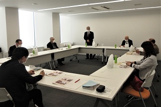 白を基調とした会議室で、ロの字型に配置されたテーブルを囲み、資料を手元に置いた参加者たちが、立って発言する男性の話を聞いている、会議風景の写真