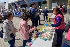 屋外のイベント会場に設置された長机で、ピンク色のボランティアベストを着用した女性スタッフが、トレイに入った飲食物をウォーキング参加者と思われる女性たちに笑顔で手渡している写真