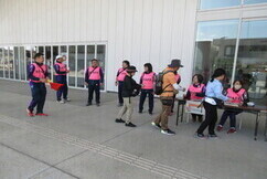 建物の軒下にある屋外の受付場所で、ピンク色のスタッフベストを着用した人々が、ウォーキングイベントの参加者と思われるリュックを背負った参加者たちの受付対応や誘導を行っている、大会運営の様子を記録した写真