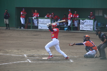 赤いユニフォームと白いズボンを着用した右打ちのバッターが、キャッチャーと審判がいるホームベースで、バットをふった瞬間の写真