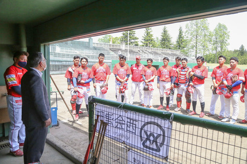 野球場のベンチ内に立ったスーツ姿の橋本市長が、グラウンドに立つ赤いユニフォームを着た野球チームのメンバーたちに向かって話している様子を捉えた写真