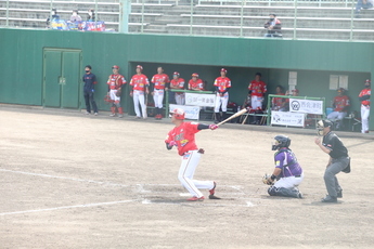 赤いユニフォームに白いズボン姿のバッターが、ピッチャー方向へ力強くバットを振りぬいた瞬間の写真