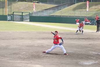 野球場のマウンド上で、赤いユニフォームと白いズボンを着用した右投げの投手が、大きく振りかぶってボールを投げようとしている瞬間の写真