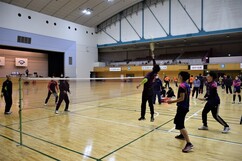 天井の高い広々とした体育館にて、ネット越しにバドミントンの三人対三人の試合が行われており、黒いユニフォームを着た選手が力強くジャンプしてシャトルを打つ瞬間を、周囲の観客や他の選手たちが見守っている風景の写真