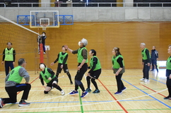 体育館にて緑色のビブスを着用したシニア世代の男女混合チームがラバーバレーボールの試合を行っており、ネット際でボールを打ち返そうとする選手や周囲で構える仲間たちの躍動感あふれる様子を捉えたスポーツ風景の写真