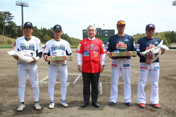 グラウンドの得点版を背景に、白いユニフォームを着た野球選手2名と濃紺のユニフォームを着た野球選手2名が、スーツの上に赤い「RED HOPES」のユニフォームを着た橋本克也市長を中央に、米袋とキュウリの箱を持って並んで立った集合写真