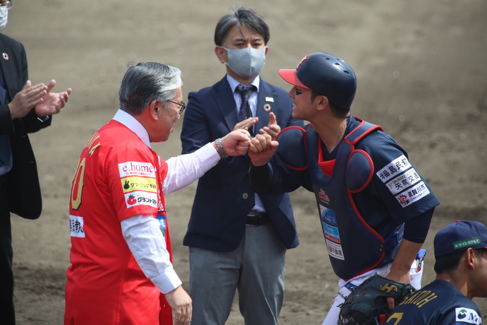 赤いユニフォームの橋本市長が、濃紺のキャッチャー防具を着用した選手とグラウンド上で拳を突き合わせている様子を、スーツ姿の男性が拍手をしながら見守っている写真