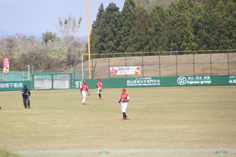 遠くに森を背景にして、立つ緑のフェンスに横断幕が設置されており、野球場の外野芝生の上に、赤いユニフォームと白いズボンを着用した3人の選手が立っている写真