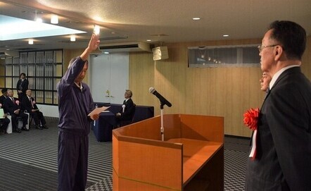 屋内の式典会場にて、紺色のトレーニングウェアを着た男性が演台の前で右手を高く掲げ、宣誓文を手に持ちながら、胸に赤いリボンをつけたスーツ姿の役員に対して力強く選手宣誓を行っている様子を横から捉えた写真