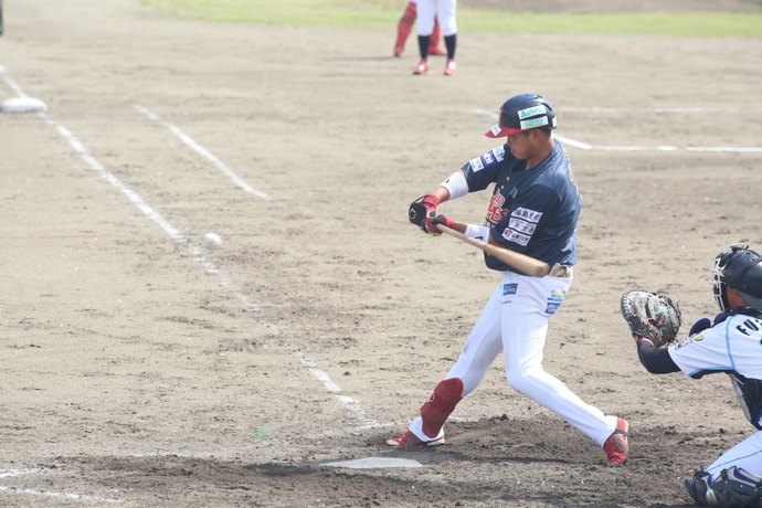 屋外の土のグランドが広がる野球場でピッチャーが投げたボールに対してバットを振り抜く紺色のユニフォーム姿の選手と、その後ろで構えるキャッチャーの写真