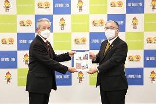 須賀川市のバックパネルの前で、マスクを着用したスーツ姿の男性二人が並び、資料を中央で一緒に持ちながら正面を向いて記念撮影を行っている写真