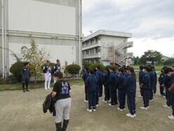 グラウンドに集合したジャージ姿の生徒たちの前で、校舎を背景にレッドホープスの選手2名と関係者の男性が立っている様子の写真