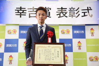 スーツ姿の相澤選手が、ステージ上の「幸吉賞 表彰式」と書かれた横断幕と須賀川市のバックパネルの前に立ち、両手に賞状とトロフィーを持っている表彰式の記念写真