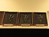 濃い茶色で、走っている人のレリーフが施された四角形の盾が3つ並んで展示されている写真