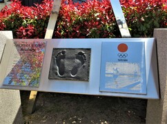 赤い葉の生垣を背景に、中央の足型のレリーフ、左側の紹介パネル、右側の五輪マークとマラソン競技の写真が配置された、円谷幸吉選手の功績を称えるステンレス製の記念碑を正面から捉えた写真