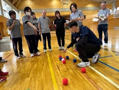体育館のコート上で、ジャージ姿の男性が床に集まった赤と青のボールを指差してルールや得点の解説を行い、その周囲を囲むように集まった7名の男女の参加者たちが熱心にその説明を聞き入っているボッチャ講習会の写真