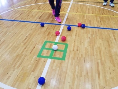 体育館の木製コートの中央に、緑色のテープで描かれた四角いターゲットマークがあり、その中心に置かれた白いボールを囲むように赤と青のボールが複数配置されている、ボッチャの競技状況を真上から捉えた写真