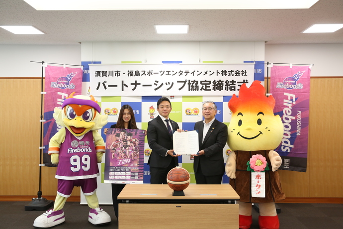 「須賀川市・福島スポーツエンタテインメント株式会社 パートナーシップ協定締結式」の横断幕と福島ファイヤーボンズののぼりの前に、紫のユニフォームを着た鳥のマスコット、福島ファイヤーボンズのポスターを持った女性、協定書を左右から持ったスーツの男性と橋本市長、ボータンのマスコットが並び、手前にバスケットボールが乗った台を置いて撮影した記念写真