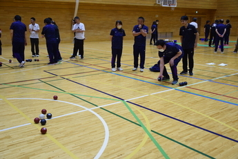 体育館のコートでニフォーム姿のたくさんの参加者たちが複数のグループに分かれてボッチャを楽しんでおり、数人の参加者が後ろに立ち見守る中、一人の男性がかがんでボールを投げようとしている様子の写真
