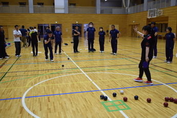 体育館のコートの周りにユニフォームを着た参加者たちが複数見守っており、たくさんのボールが集まっているコートの円の中を目掛けて、男性がボールを投げようとしている様子の写真
